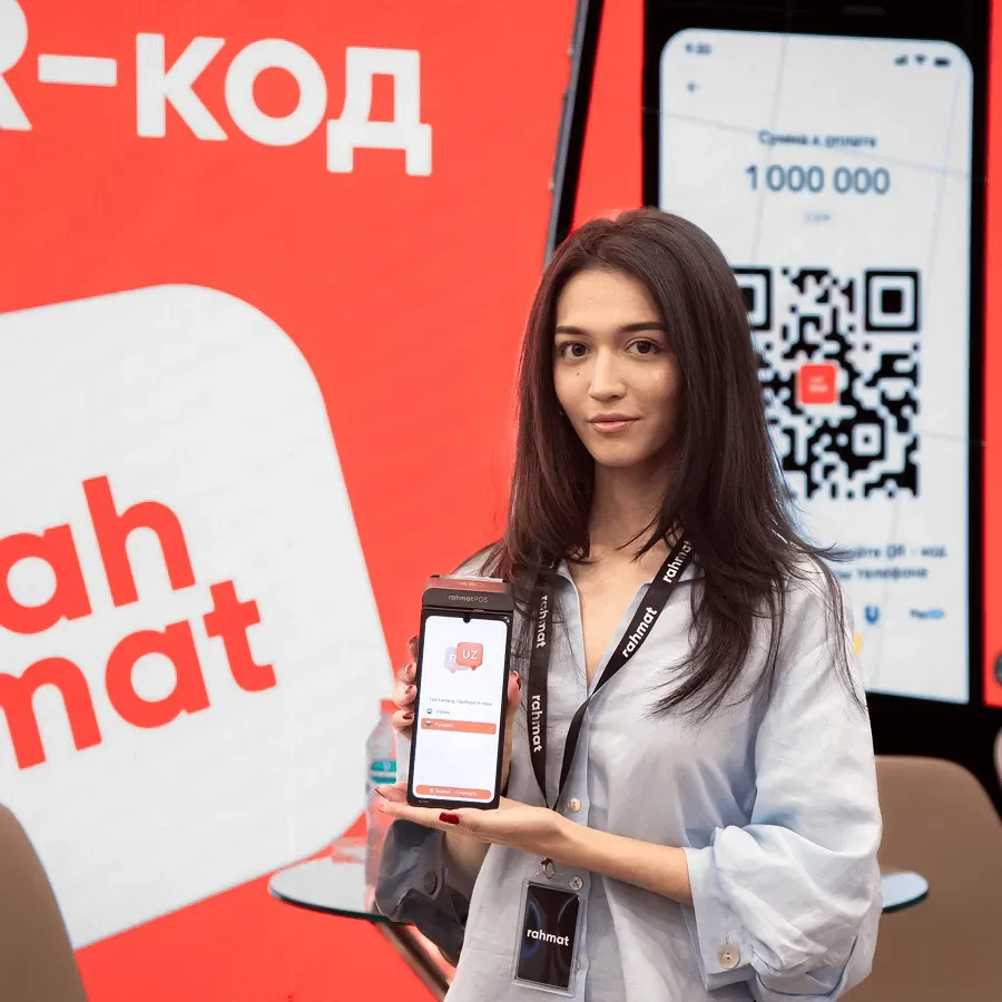 A Rahmat employee holding a Rahmat POS terminal in front of a QR code screen